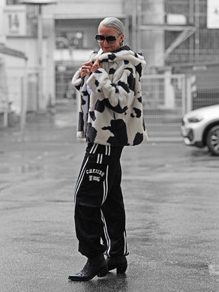 Person trägt OFF#DLY Joggpant Giorgia black mit CHERISH und 15-Aufdruck, schmalem Schnitt und elastischem Bund, kombiniert mit schwarzen Stiefeln und einer gepunkteten Fake-Fur-Jacke bei nassem Wetter.