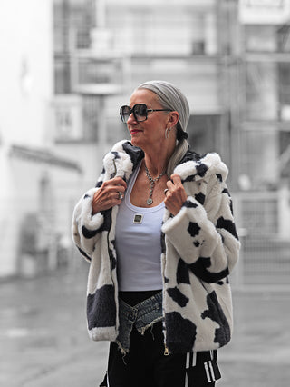 Frau trägt OFF#DLY Fake Fur Jacke Animal black/white mit auffälligem Kuhmuster, Kapuze und Reißverschluss. Sie hält den Kragen und blickt lächelnd weg, kombiniert mit sportlicher Hose und Sonnenbrille.