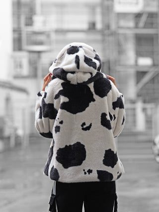 Person von hinten auf einer Straße, trägt eine OFF#DLY Fake Fur Jacke Animal black/white mit Kapuze und auffälligem schwarz-weißen Kuhmuster, hält die Hände am Kragen, Hintergrund unscharf.
