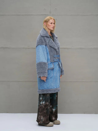 #VDR Mantel 18077 blue: Frau im Profil trägt einen mittelblauen Patchwork-Denimmantel mit grauem Faux-Fur-Besatz an Kragen, Ärmeln und Saum, Taillengürtel und großen Fronttaschen.