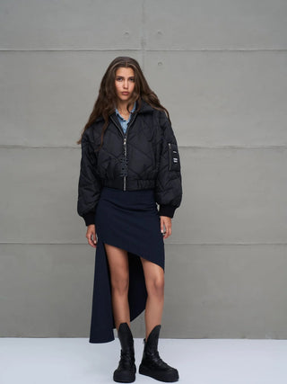 Junge Frau trägt die #VDR Jacket 18138 black, eine kurze, gesteppte Bomberjacke mit elastischem Bund und Frontzipper, kombiniert mit asymmetrischem Rock und klobigen Stiefeln, vor grauer Wand.