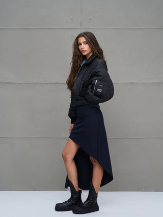 Junge Frau im Profil trägt #VDR Jacket 18138 black, eine kurze, gesteppte Bomberjacke mit Frontzipper und elastischem Bund, kombiniert mit asymmetrischem Rock und klobigen schwarzen Stiefeln.