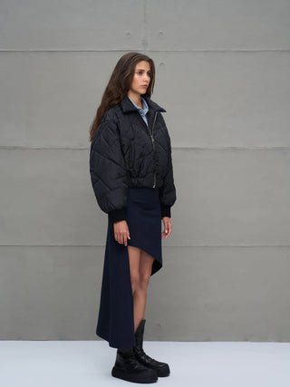 Junge Frau im Profil trägt #VDR Jacket 18138 black, eine kurze, gesteppte Bomberjacke mit Frontreißverschluss und elastischen Bündchen, über einem Denimhemd mit asymmetrischem Rock und Plateau-Stiefeln.