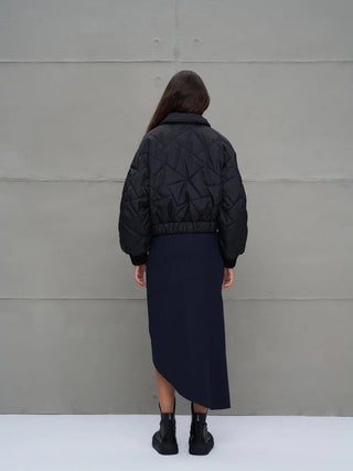 Person von hinten vor grauer Wand, trägt #VDR Jacket 18138 black. Die schwarze, gesteppte, kurze Bomberjacke hat Puffärmel und betont durch elastischen Bund die Silhouette, kombiniert mit asymmetrischem Rock und Stiefeln.