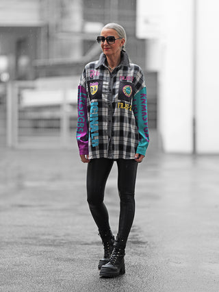 Frau trägt die OFF#DLY Checker Bluse Frida black/grey über glänzenden Leggings, dazu klobige schwarze Stiefel. Die Bluse zeigt ein kariertes Muster mit bunten Patches und Schriftzügen an Ärmeln und Brust.