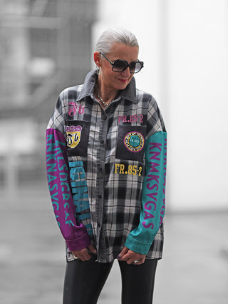 Ältere Frau trägt die OFF#DLY Checker Bluse Frida black/grey, ein oversized kariertes Hemd mit grafischen Patches und Schriftzügen, kombiniert mit schwarzen Leggings.