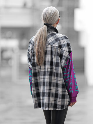 Person von hinten mit langem grauweißem Haar trägt die OFF#DLY Checker Bluse Frida black/grey, ein schwarz-weiß kariertes Hemd mit lila und türkis bedruckten Ärmeln, über einem lila Langarmshirt.