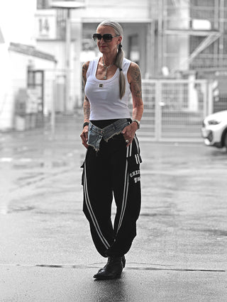 Frau trägt OFF#DLY Joggpant Giorgia black mit weißen Streifen und CHE-Logo, kombiniert mit weißem Tanktop und Denim-Detail. Tattoos sichtbar, Accessoires und sporty urbaner Stil unterstreichen den Look.