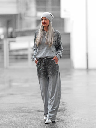 Ältere Frau in OFF#DLY Jogginganzug BlingBling grey, bestehend aus lockerem Hoodie und passenden Hosen mit Strassdetails, trägt eine graue Beanie, steht lässig auf nassem Stadtpflaster.