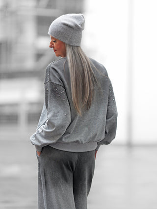 Frau von hinten in urbaner Umgebung trägt den OFF#DLY Jogginganzug BlingBling grey mit Strass-Details auf grauem Sweatshirt und Hose, ergänzt durch graue Beanie, Hände in Taschen.