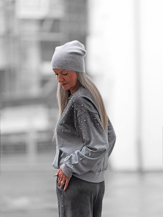 Ältere Frau trägt den OFF#DLY Jogginganzug BlingBling grey mit Strass-Details, bestehend aus grauem Beanie, Sweatshirt und Sweatpants, bei Regen in urbaner Umgebung.