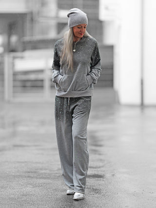 Person in OFF#DLY Jogginganzug BlingBling grey, bestehend aus lockerem, strassbesetztem Sweatshirt und Sweatpants mit Kängurutasche, spaziert auf nassem Gehweg.