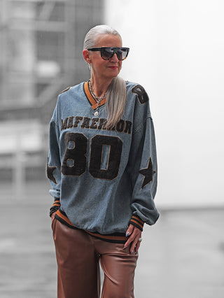 Silberhaarige Frau trägt das OFF#DLY Denimshirt Ariane blue mit „80“-Print und Stern-Applikationen, kombiniert mit hohen braunen Lederhosen, zeigt lässigen und sportlichen Vintage-Stil.