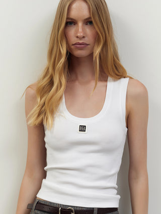 Junge Frau trägt BSB Top 210020 white, ein figurbetontes, ärmelloses Tanktop mit Rippstruktur und kleinem Logo “ESS3” auf der Brust, kombiniert mit karierten Hosen und dunkelbraunem Ledergürtel.