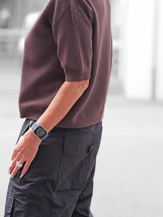 Person mit dem OFF#DLY Pulli Tina schoko, einem kurzärmeligen, eng geschnittenen Stricktop, kombiniert mit lockeren schwarzen Cargohosen und einer quadratischen Smartwatch am Handgelenk.