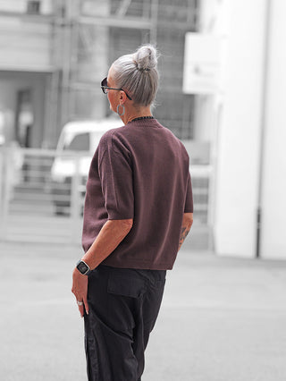 Person mit silbernem Dutt trägt OFF#DLY Pulli Tina schoko, kurzärmelig und kastig. Dazu schwarze Cargo-Hose, Sonnenbrille, geometrischer Ohrring und Smartwatch. Urbaner Hintergrund mit unscharfen Gebäuden.