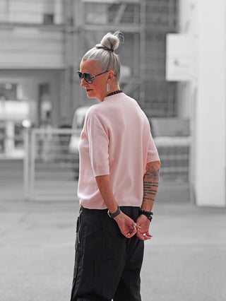 Person mit silbergrauem Haar im Dutt trägt den OFF#DLY Pulli Tina rosa, einen schmal geschnittenen, kurzärmeligen Pullover, kombiniert mit schwarzen Cargo-Hosen, Sonnenbrille und auffälligem Schmuck.