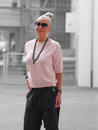 Ältere Frau trägt den OFF#DLY Pulli Tina rosa, ein kurzärmliges, schmal geschnittenes, rosafarbenes Oberteil, kombiniert mit schwarzen Cargo-Hosen und Accessoires, darunter eine lange Kette und eine Uhr.