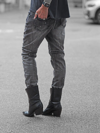 Person trägt OFF#DLY Denim Ostia: Slim-fit, charcoal-black Jeans mit weißen Farbsprenkeln, kombiniert mit glatten schwarzen Lederstiefeln. Hand mit Ringen und Smartwatch sichtbar. Perfekt für modernen Streetstyle.