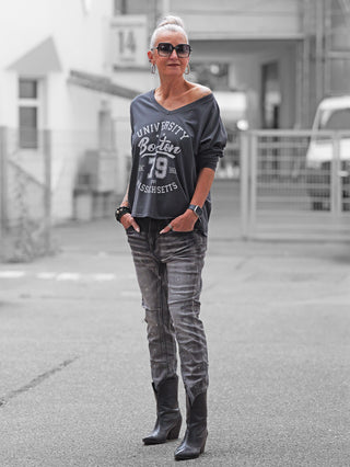 Ältere Frau trägt OFF#DLY Denim Ostia, lockere Jeans mit Used-Effekten und Farbsprenkeln, kombiniert mit schwarzem Sweatshirt und eleganten Ankle Boots, perfekt für modernen Streetstyle.