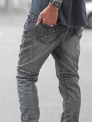 Nahaufnahme der OFF#DLY Denim Ostia Jeans mit schmaler Passform, dezenten Farbsprenkeln und Knie-Nähten. Die rechte Hand mit Ringen steckt in der Gesäßtasche, links eine dunkle Smartwatch.
