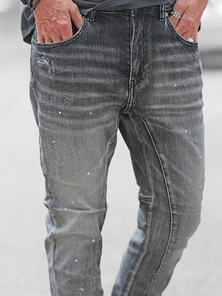 Nahaufnahme von der unteren Körperhälfte einer Person, die OFF#DLY Denim Ostia trägt. Die Jeans in verwaschenem Schwarz zeigt dezente Farbsprenkler und Used-Effekte, Hände in den Taschen, mit Ringen und Armband.