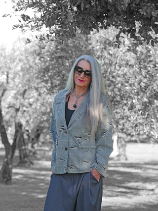 Person in einem OFF#DLY Blazer Arabba Checker mit klassischem Karomuster und Destroyed-Details, trägt Sonnenbrille, pinken Lippenstift, schwarze Hose, steht lässig unter Baum.