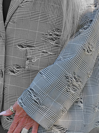 OFF#DLY Blazer Arabba Checker, ein stark zerstörter Hahnentritt-Blazer mit Fransen, getragen über einem schwarzen Top. Der Träger zeigt silberne Ringe und helle, lange Nägel.