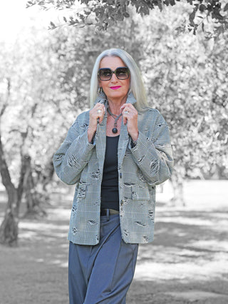 Ältere Frau im Park trägt den OFF#DLY Blazer Arabba Checker mit klassischem Karomuster und Destroyed-Details, kombiniert mit grauer Hose. Sie hält die Blazer-Lapels, betont durch Sonnenbrille und Schmuck.