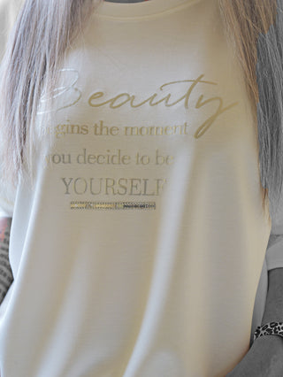 Person trägt OFF#DLY Sweater Beauty black white mit dem Satz „Beauty begins the moment you decide to be YOURSELF“ in Gold, funkelndes Detail darunter, lässiger Schnitt, sichtbares Tattoo.