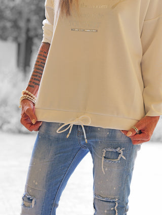 Person trägt OFF#DLY Sweater Beauty black white mit lockerem Schnitt und Statement-Print, kombiniert mit hellblauen, zerrissenen Jeans und Perlarmbändern, betont durch neutralen Nagellack.