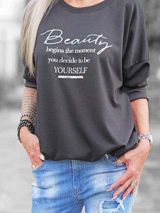 Eine Person trägt den OFF#DLY Sweater Beauty black mit weißem Statement-Print „Beauty begins the moment you decide to be yourself“, kombiniert mit hellen, leicht zerrissenen Jeans und auffälligen Accessoires.