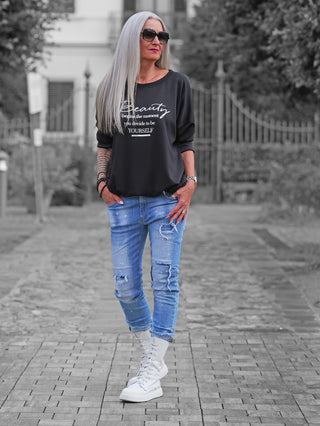 Frau trägt den OFF#DLY Sweater Beauty black mit inspirierendem Print, kombiniert mit hellblauen Jeans und weißen Stiefeln, während sie einen lässigen und selbstbewussten Stil in einer Gartenumgebung zeigt.