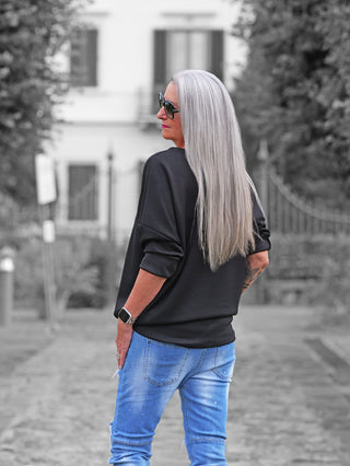 Eine Frau mit silber-grauen Haaren trägt den OFF#DLY Sweater Beauty black mit lässigem Schnitt und Statement-Print. Kombiniert mit blauen Jeans, zeigt sie einen modernen, komfortablen Stil.