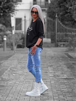 Person trägt OFF#DLY Sweater Beauty black, einen locker geschnittenen schwarzen Pullover mit weißem Statement-Print. Kombiniert mit hellblauen Jeans und weißen Stiefeln auf einem gepflasterten Weg.