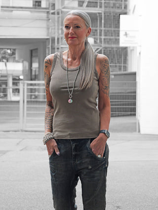 Mittelalte Person trägt das OFF#DLY Top Neve taupe, ein lässiges ärmelloses Baumwoll-Top mit Smiley-Patch, kombiniert mit dunklen Jeans in urbaner Umgebung.