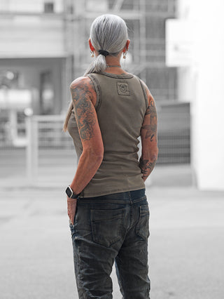 Person von hinten in urbaner Umgebung trägt ein ärmelloses OFF#DLY Top Neve taupe mit Smiley-Patch am Rücken, kombiniert mit dunklen Jeans und einer Smartwatch am linken Handgelenk.