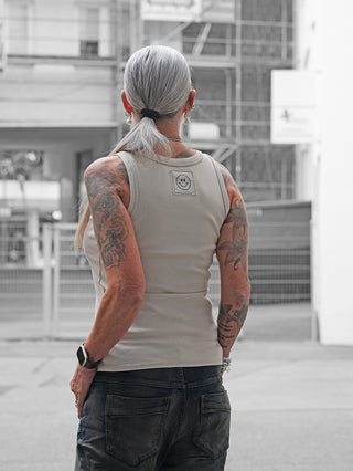 Person von hinten mit dem OFF#DLY Top Neve beige, ärmellos mit Smiley-Patch auf dem oberen Rücken, kombiniert mit dunklen Jeans. Silbergrauer Pferdeschwanz, Tattoos, und dezentem Schmuck sichtbar.