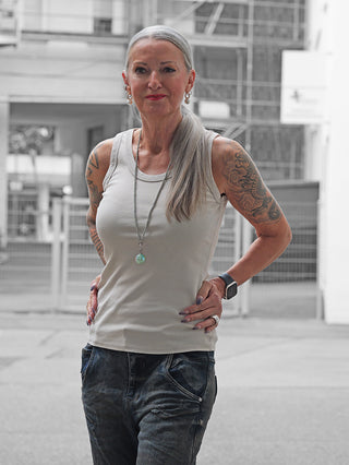 Ältere Frau trägt das OFF#DLY Top Neve beige, ein eng anliegendes Tanktop mit einem Smiley-Patch, kombiniert mit dunklen Jeans, Schmuck und sichtbaren Tattoos in urbaner Umgebung.