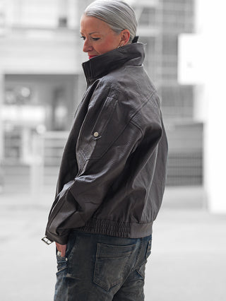 Frau in urbaner Umgebung trägt die OFF#DLY Biker Jacke coffee, oversized mit hochgestelltem Kragen, kombiniert mit Jeans. Jacke hat markante Struktur und Zipper-Details, strahlt urbanen Charme aus.