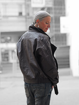 Person im Profil trägt die OFF#DLY Biker Jacke coffee, eine oversized Lederjacke mit Reißverschluss-Details und extragroßen Revers, kombiniert mit Jeans und auffälligem Perlen-Ohrring, in urbanem Umfeld.