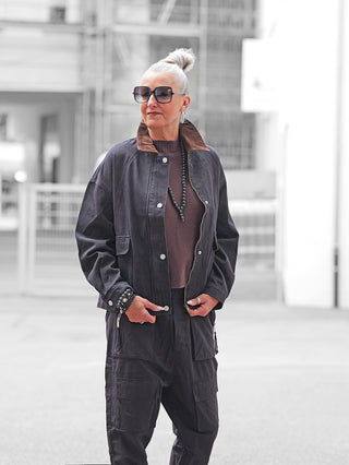 Ältere Frau in urbaner Umgebung trägt die OFF#DLY Jacke Denim Black, eine oversized Denimjacke mit braunem Cordkragen, über einem braunen Pullover, kombiniert mit schwarzen Cargohosen und Accessoires.