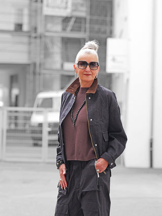 Frau mit silbernem Haar trägt die OFF#DLY Jacke Denim Black über braunem Pullover, kombiniert mit schwarzen Cargo-Hosen, Accessoires und Sonnenbrille, in urbaner Umgebung stehend, wirkt selbstbewusst in die Kamera blickend.