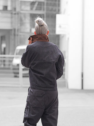 Eine Person mit silbernem Haarknoten trägt die OFF#DLY Jacke Denim Black, eine übergroße, dunkle Jacke mit hochgeklapptem Kragen, passend zu lässig geschnittenen Cargo-Hosen, in urbaner Umgebung.