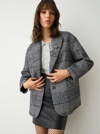 Junge Frau trägt BSB Blazer Checker carbon mit markantem Karo-Muster, dekorativer Brosche am Revers, über grauem Strickpullover und schwarzem Lederrock, kombiniert mit Netzstrümpfen.