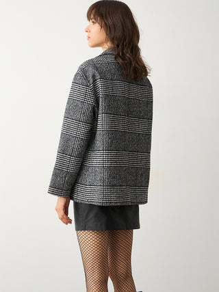 Frau trägt BSB Blazer Checker carbon, ein karierter Glencheck-Blazer mit dekorativer Brosche, über kurzem Lederrock und Netzstrumpfhose, von hinten in stilvoller Pose gegen einen hellen Hintergrund.