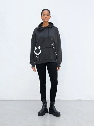 Das Model trägt den Outryght Sweater HAZ S. in verwaschenem Schwarz mit einem weißen, farbgesprenkelten Smiley-Grafik, kombiniert mit engen schwarzen Jeans und klobigen schwarzen Lederstiefeln.