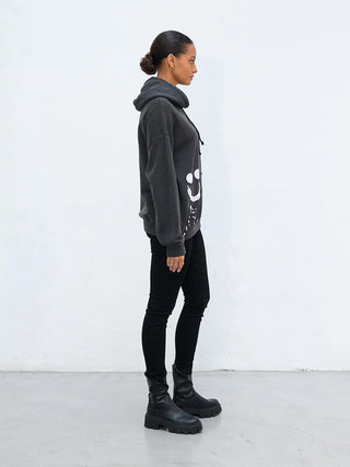 Frau in einem übergroßen Outryght Sweater HAZ S. faded black mit handgemaltem Smiley-Motiv, kombiniert mit schwarzen Skinny-Jeans und klobigen, schwarzen Stiefeletten, im Profil vor weißer Wand stehend.