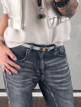 Nahaufnahme eines schlanken, hellgrauen OFF#DLY Gürtel Leder mit goldfarbener Schnalle, getragen über verblassten, schwarzgrauen Jeans. Sichtbare Accessoires umfassen eine weiße Smartwatch und schwarze Perlenarmbänder.