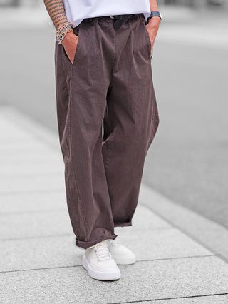 Person trägt OFF#DLY Joggpant Melania schoko, weite braune Baumwollhose mit umgeschlagenen Säumen, seitlichen Taschen, sichtbarem weißen T-Shirt und weißen Sneakers. Arm mit Kettenarmbändern und Smartwatch.